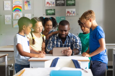 African American young male teacher showing digital tablet to multiracial elementary students
